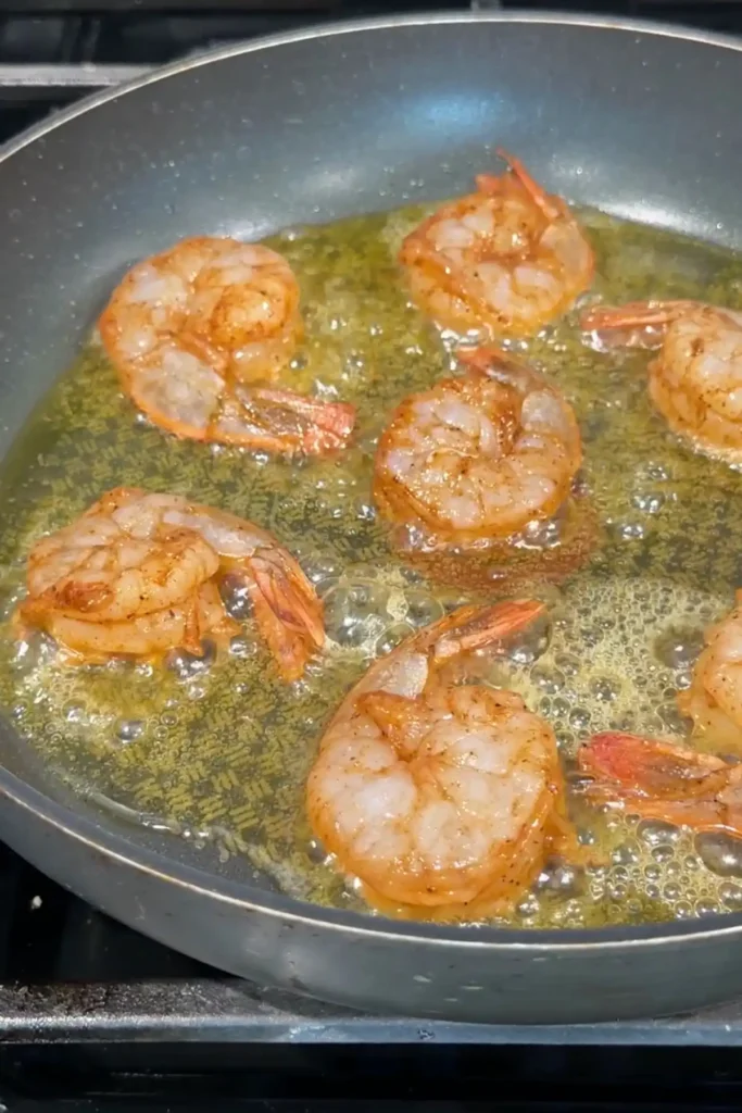 Seasoned shrimp cooking in a gray nonstick skillet with bubbling butter and oil, beginning to turn pink around the edges while searing on the stovetop.