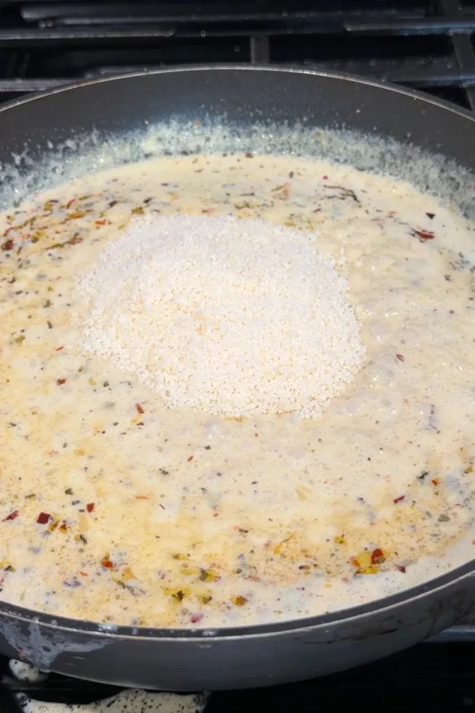 Heavy cream sauce simmering in a skillet with herbs and red pepper flakes, topped with a mound of grated parmesan in the center just before stirring.