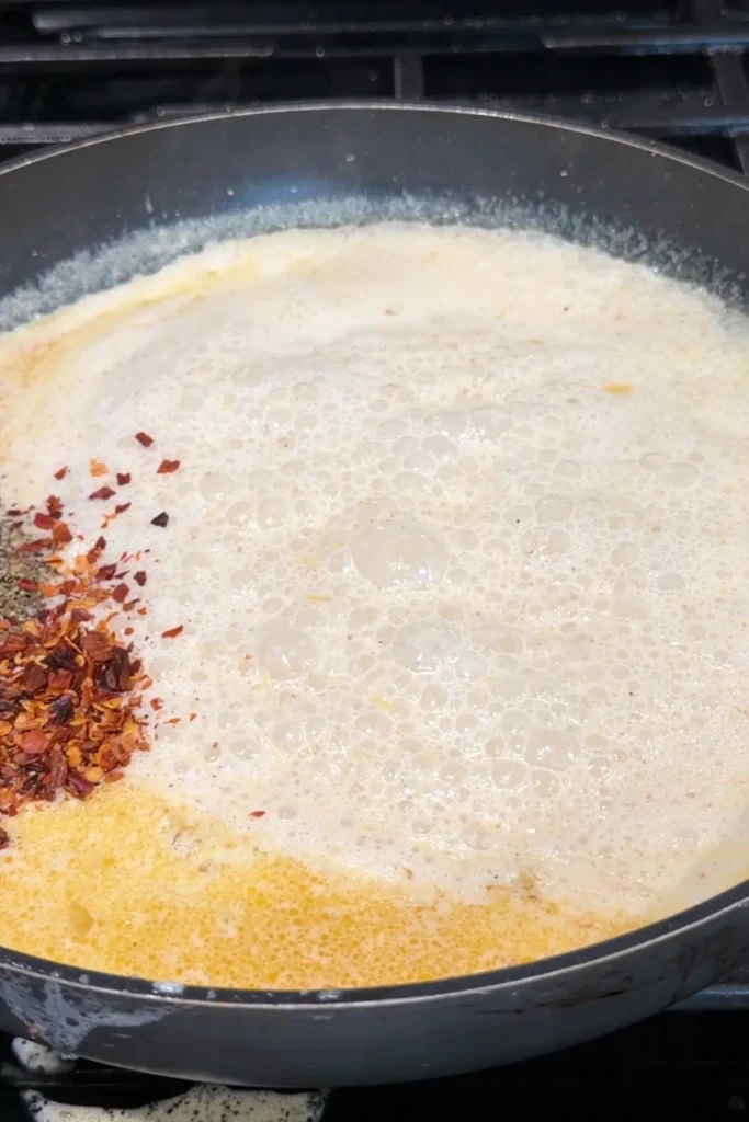 Creamy sauce bubbling in the skillet with red pepper flakes and black pepper added on one side, showing the seasonings before they are mixed in.