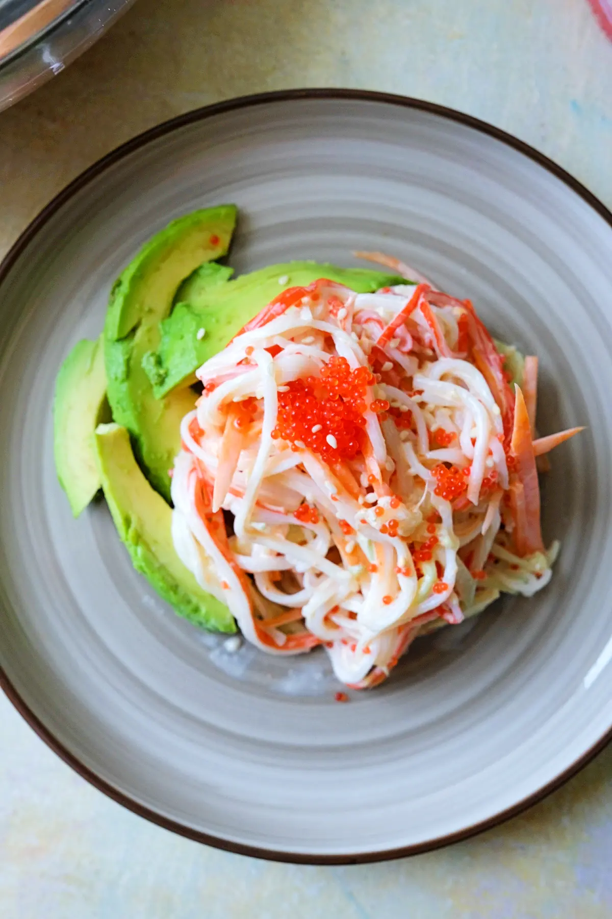 Overhead view of a plate holding a neat serving of kani salad topped with a spoonful of bright orange tobiko, with avocado slices arranged along the side.