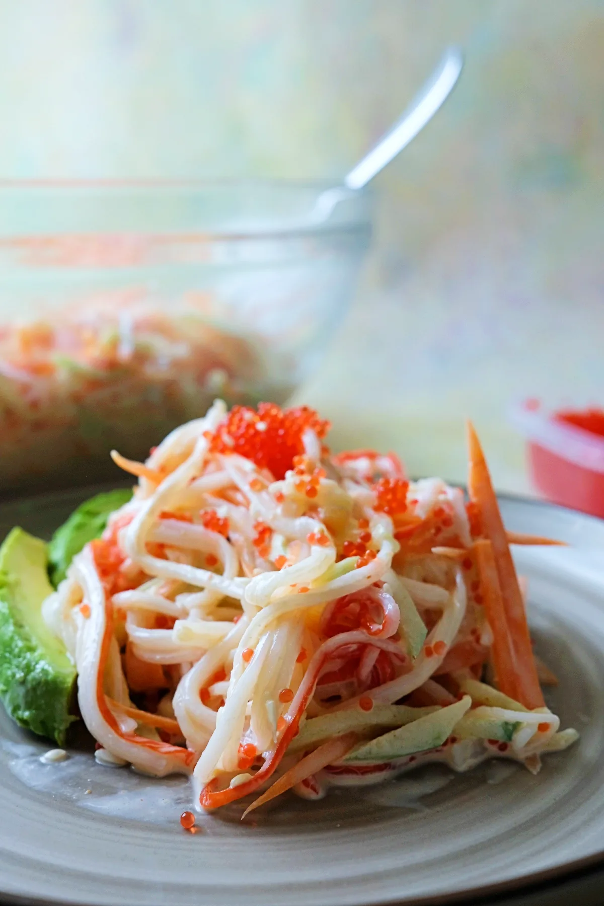 A mound of spicy kani salad made with shredded imitation crab, julienned cucumber, and carrot sits on a plate, topped with bright orange tobiko and served beside sliced avocado.
