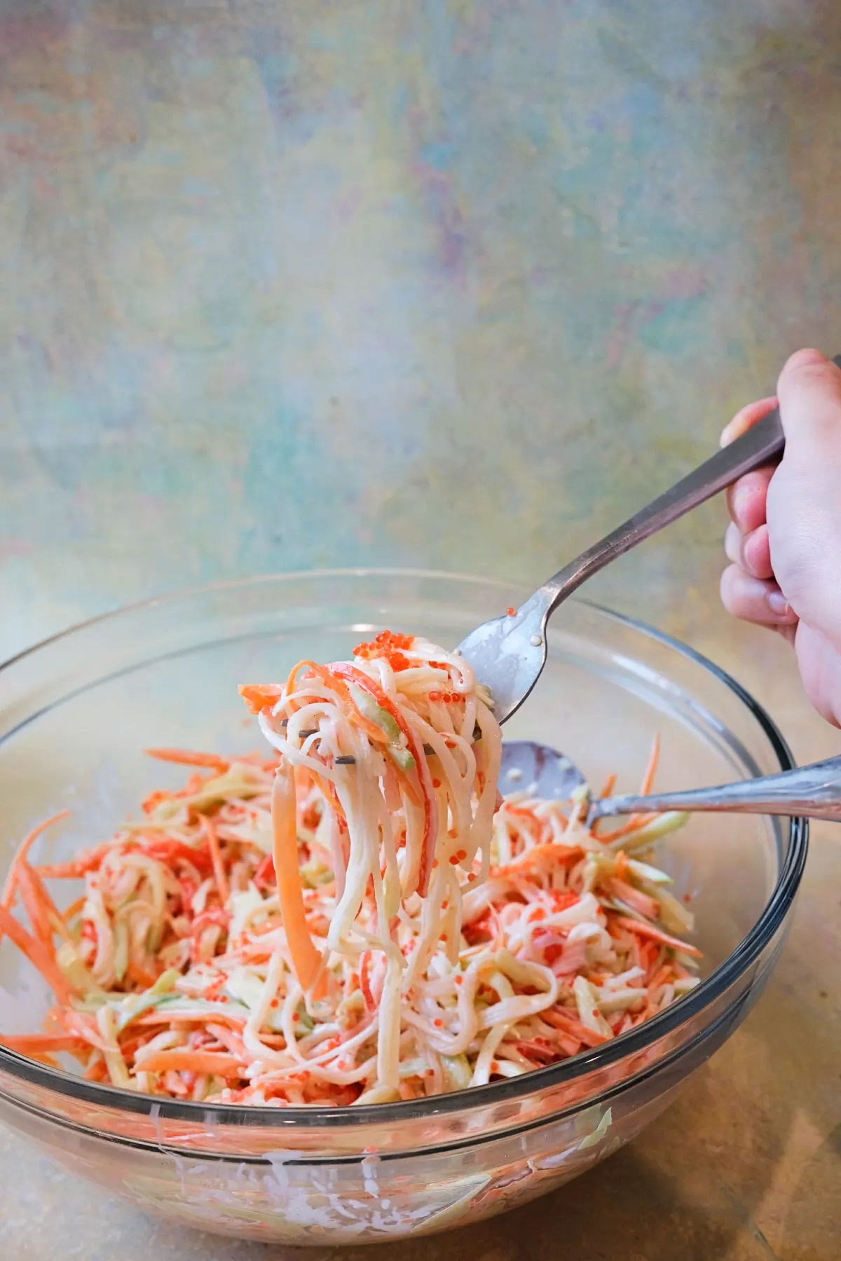 A hand lifts a forkful of kani salad from a large clear glass bowl, showing long strands of imitation crab, thin cucumber, carrot, and small orange tobiko pearls coated in creamy dressing.