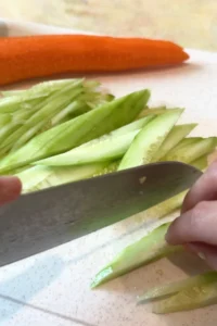 A hand uses a chef’s knife to julienne cucumber into thin matchsticks on a white cutting board