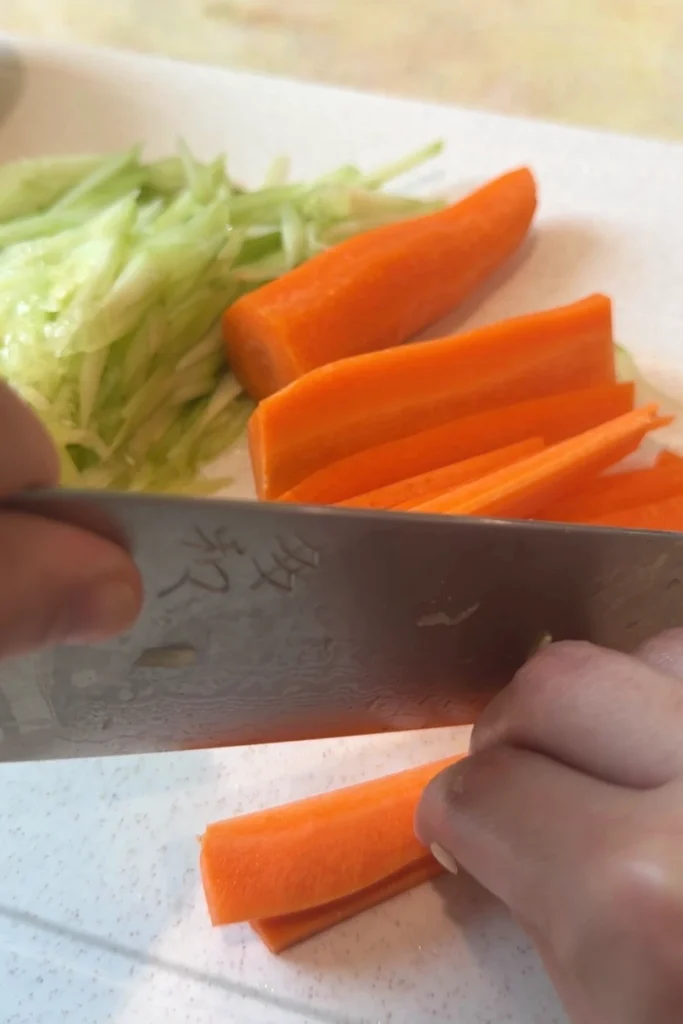 A hand slices carrot into thin planks on a white cutting board with a chef’s knife