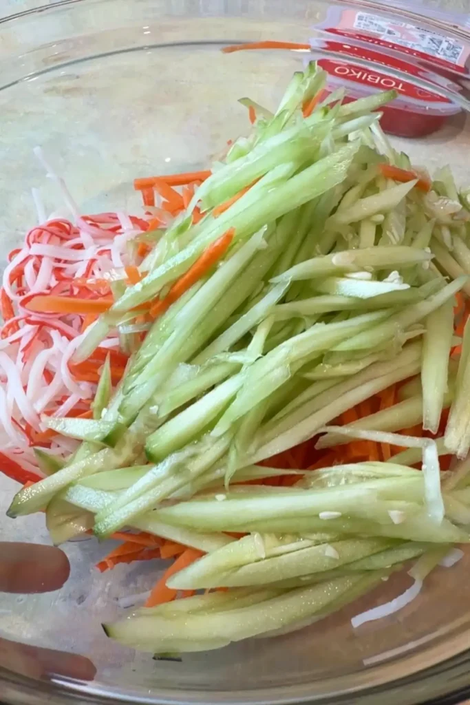 Julienned cucumber and carrot are added to a large glass bowl with shredded imitation crab, creating colorful layers of green, orange, and red-and-white strips.