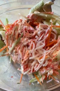 A spoon tosses the kani salad ingredients together in a glass bowl, coating the imitation crab, cucumber, and carrot in a creamy spicy dressing with tobiko mixed throughout.