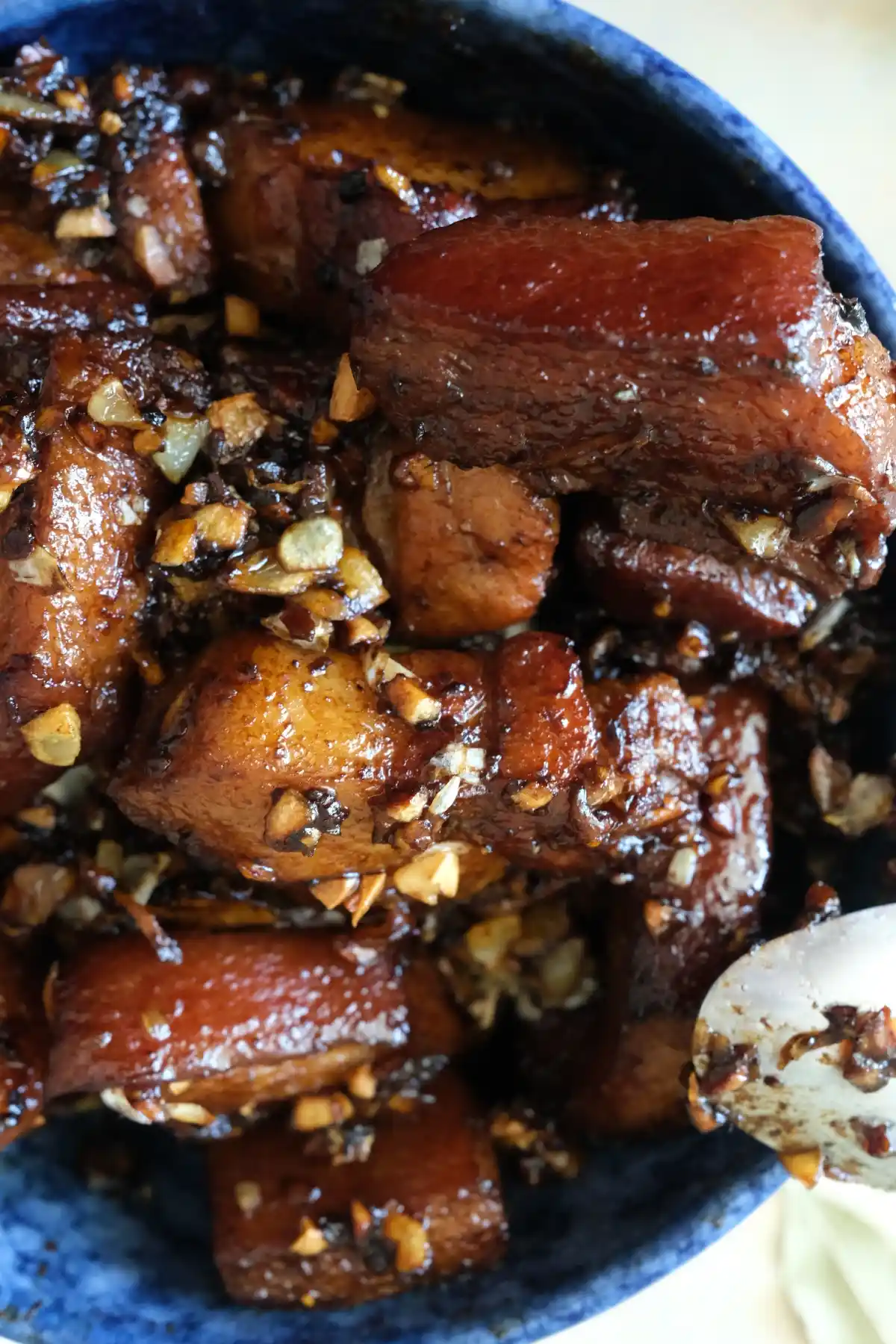 Overhead close-up of pork belly adobo in a blue bowl, showing thick, tender pieces of pork coated in a deep brown sauce. 