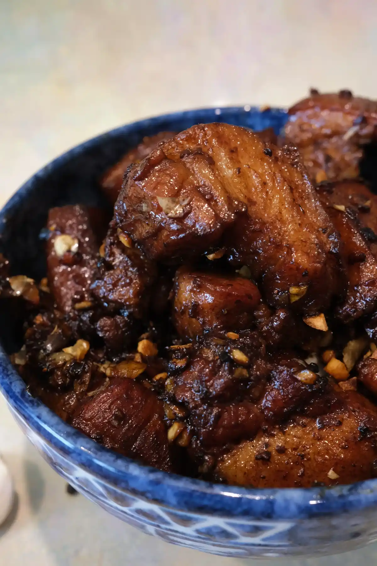 Close-up of Filipino pork adobo in a blue ceramic bowl, with glossy chunks of pork belly stacked high and coated in a dark soy-vinegar sauce.