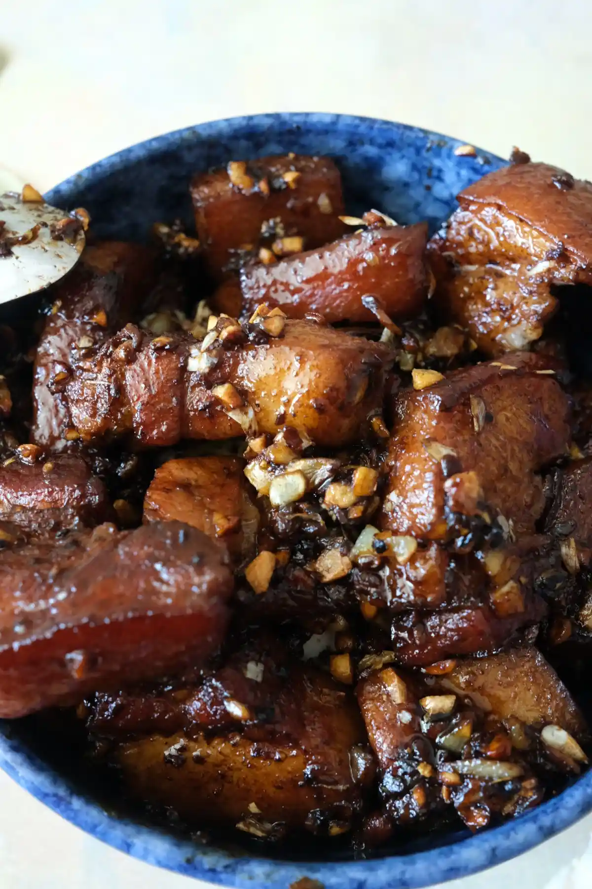 A blue bowl filled with pork adobo, with glossy pork belly pieces piled over one another and topped with crispy garlic. A bowl of white rice sits just off to the side, suggesting how the dish is served.