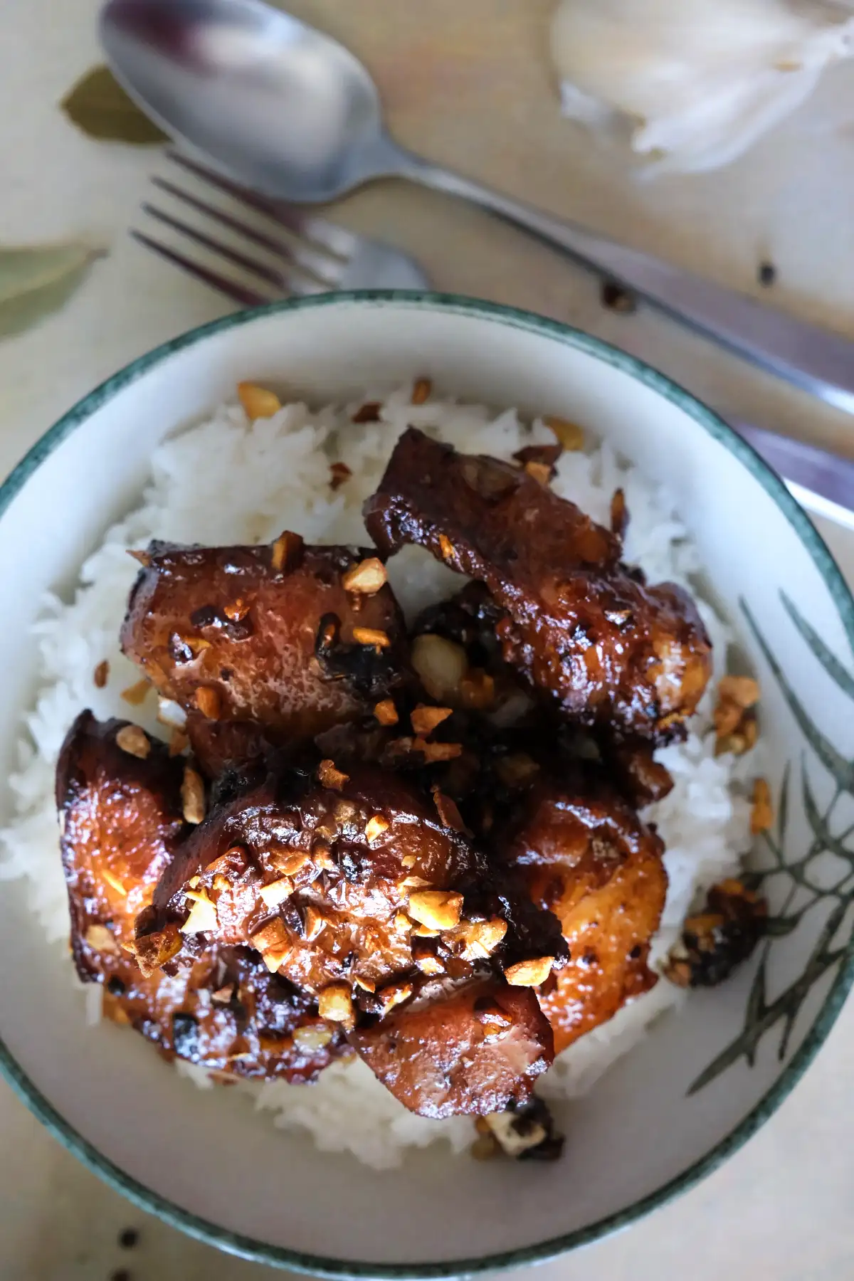 Top-down view of pork adobo served over white rice in a small ceramic bowl. The glossy pork belly is piled in the center and sprinkled with browned garlic, with a spoon and fork blurred in the background.