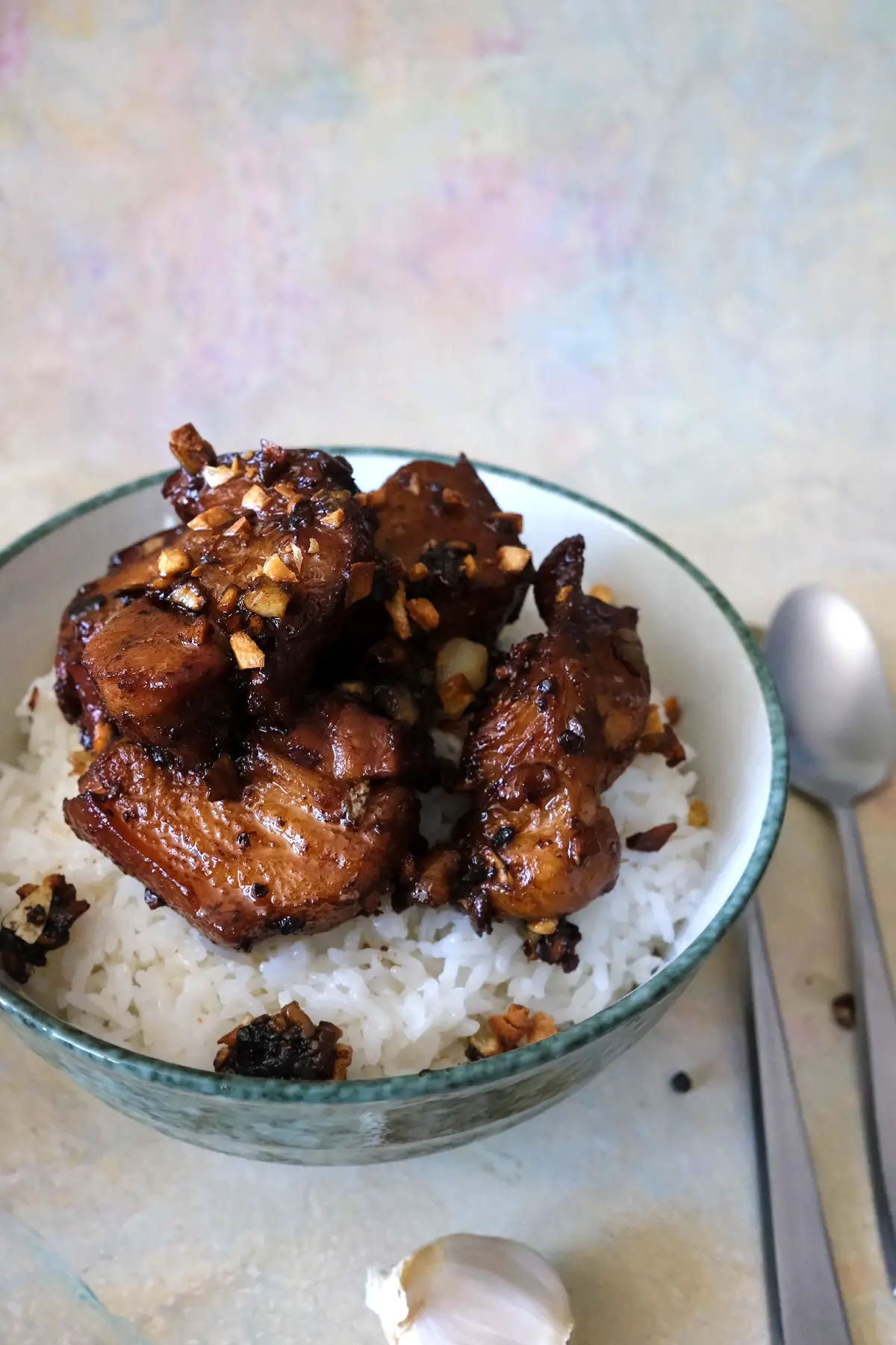 pork adobo served over white rice in a green-rimmed bowl, with chunky pork belly pieces stacked on top and finished with browned garlic. A whole garlic bulb and spoon sit nearby, tying in the garlicky flavor of the dish.