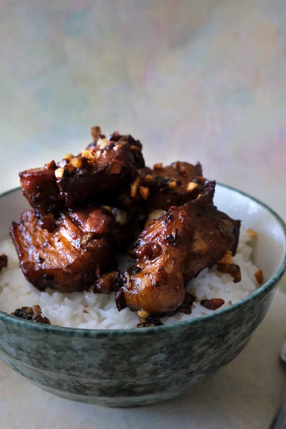 A bowl of white rice topped with rich pork adobo, showing thick, glistening pork belly pieces with caramelized edges. Crisp browned garlic is scattered across the meat and rice