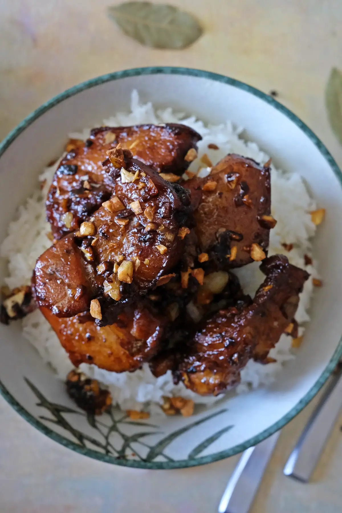 Overhead shot of a bowl of white rice topped with several pieces of pork belly adobo, each coated in a dark glossy sauce and scattered with crisp garlic. The bowl sits beside silverware, with a few browned garlic bits around the rim and table.