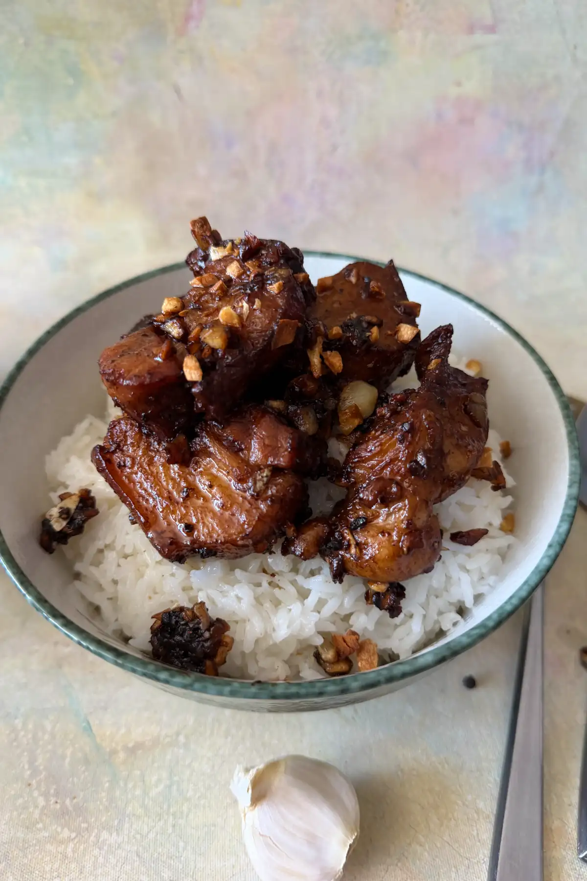 pork adobo served over white rice in a patterned bowl, with several dark, glossy pork belly pieces arranged in the center and finished with crisp garlic. Bay leaves, peppercorns, silverware, and a garlic clove are scattered around the bowl