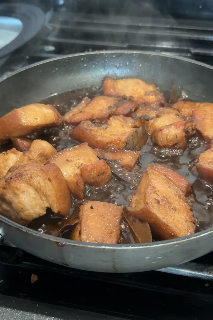 Pork belly simmering in a dark adobo sauce in a wide skillet, with bay leaves visible between the pieces.