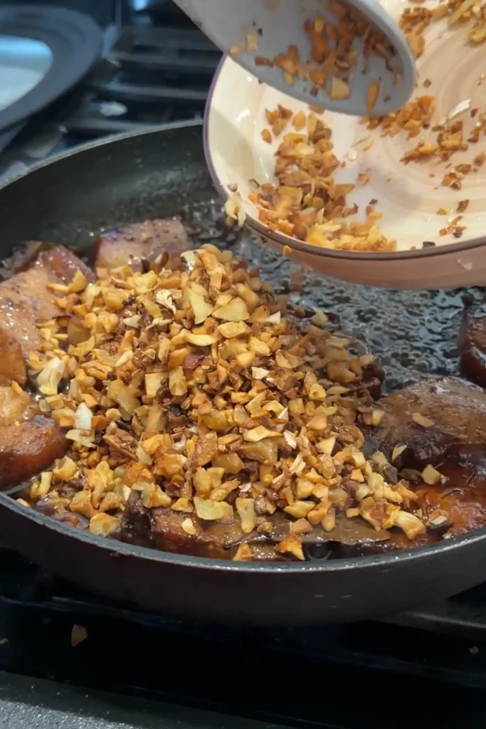 Crispy browned garlic being tipped back into the skillet over the simmering pork adobo.