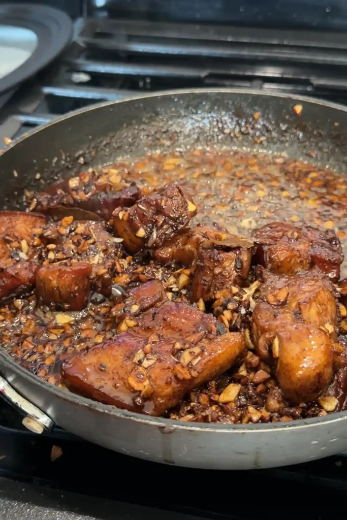 Finished pork adobo in a skillet, with glossy browned pork belly coated in a thick dark sauce and plenty of crisp garlic scattered throughout.