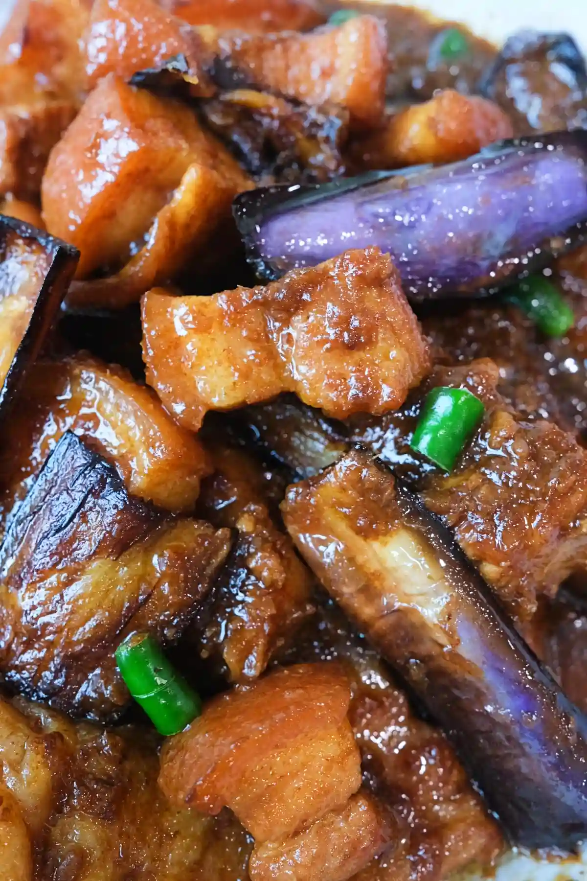 Extreme close-up of pork binagoongan with glossy pork belly cubes, fried eggplant, and sliced green chilies coated in a thick shrimp paste sauce.