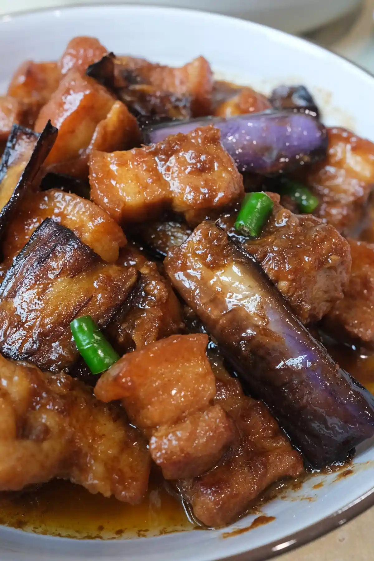 Close-up of pork binagoongan in a white bowl, showing tender pork belly, purple eggplant pieces, green chilies, and a rich brown sauce clinging to every piece.