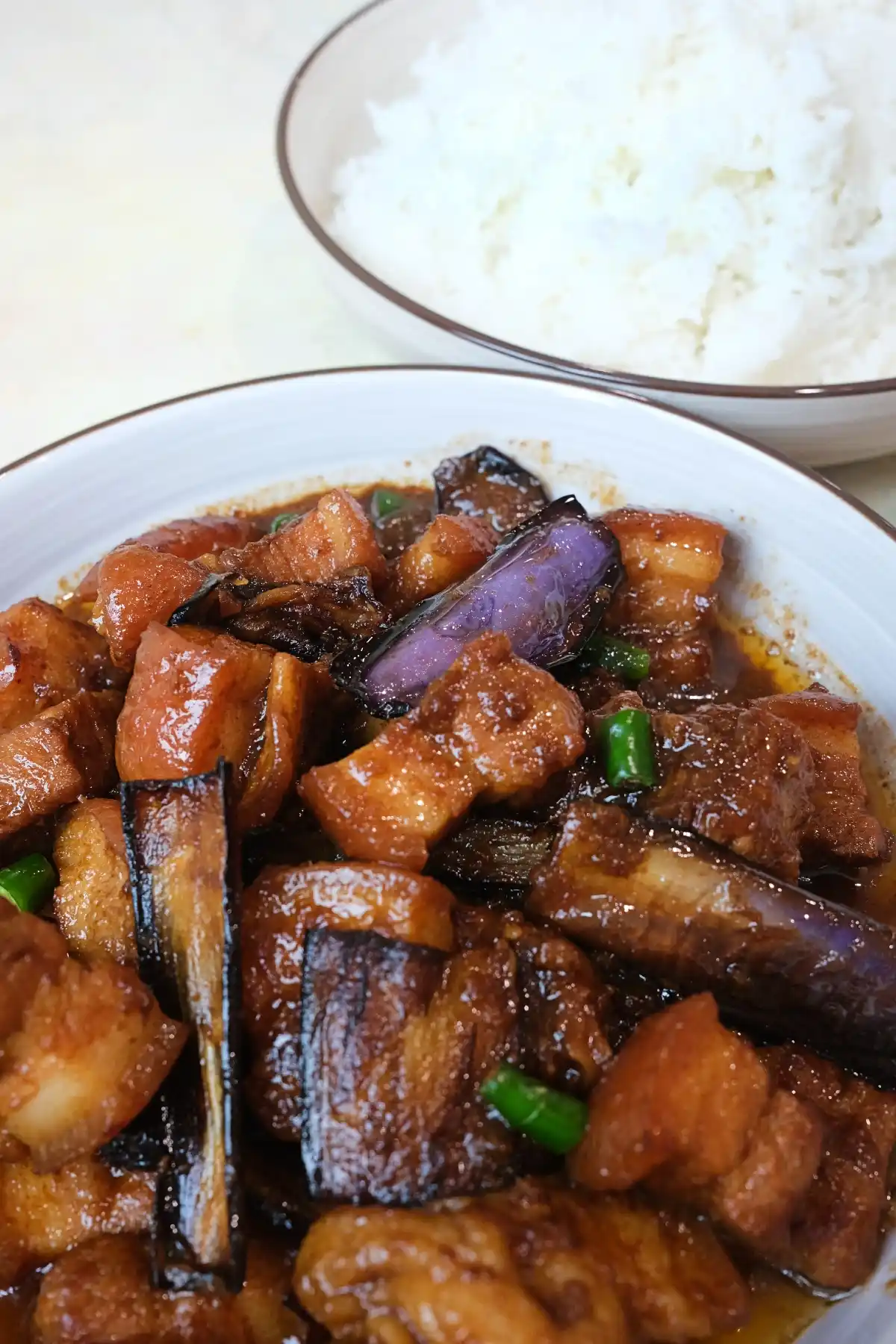 Bowl of pork binagoongan with eggplant and green chilies, served beside a separate bowl of white rice in the background.