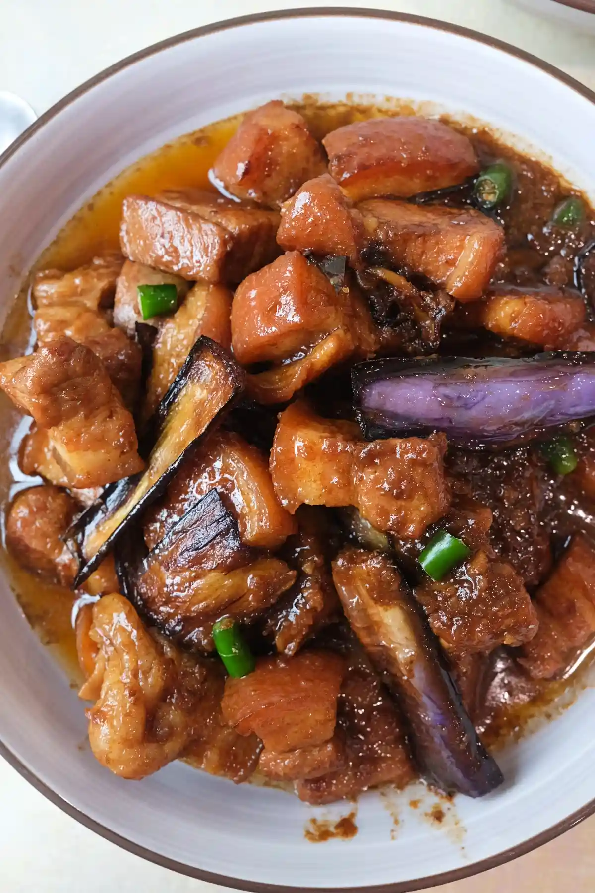 Overhead view of pork binagoongan in a shallow bowl with chunky pork belly, fried eggplant, green chilies, and a rich oily shrimp paste sauce.