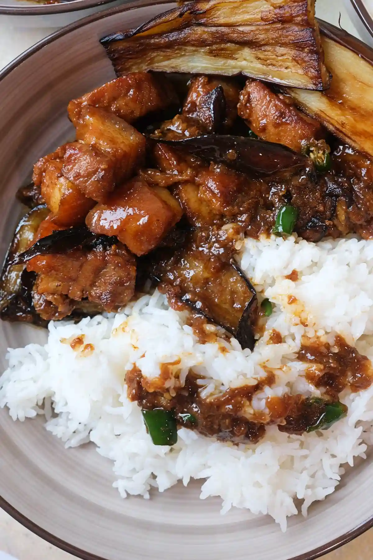 Plate of white rice topped with pork binagoongan, featuring glossy pork belly, saucy eggplant, chopped green chilies, and long fried eggplant slices on top.