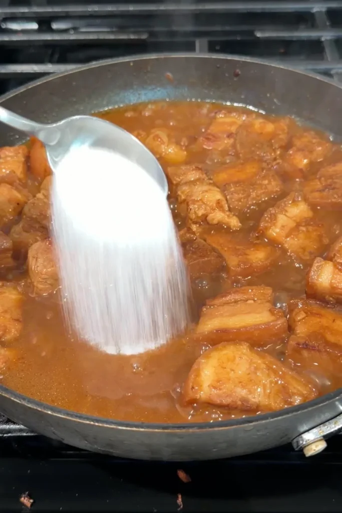 Sugar being added by spoonful to the simmering pork binagoongan sauce.