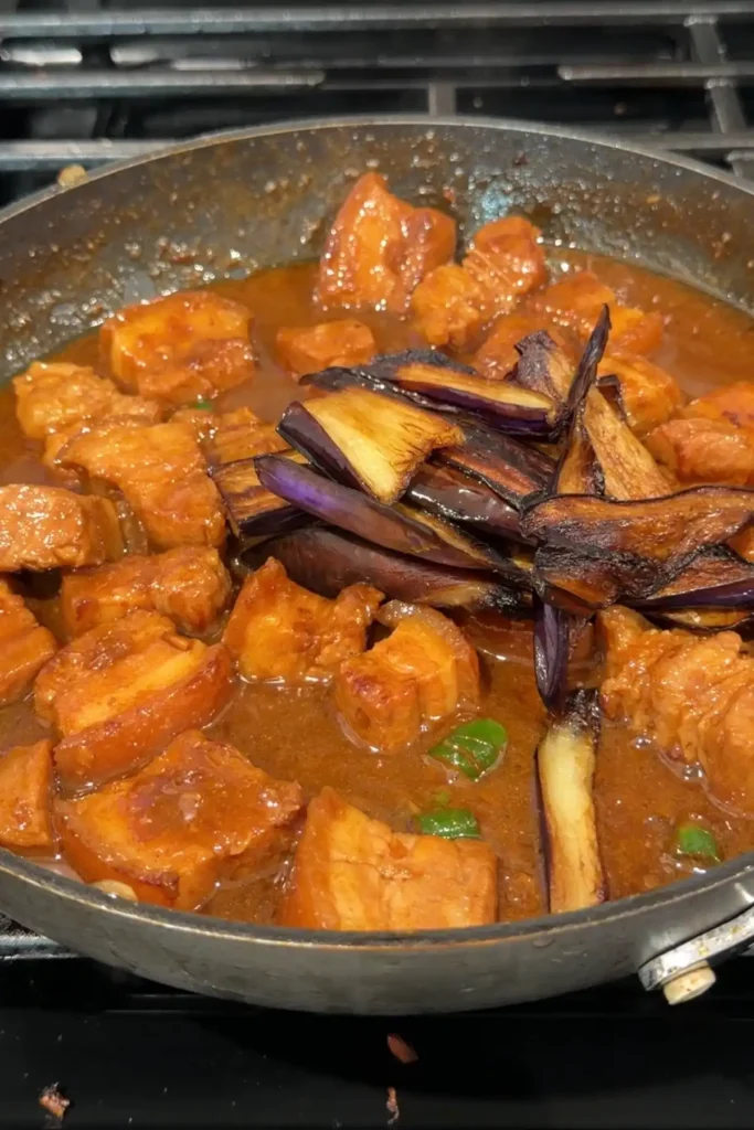 Fried eggplant being added back to the skillet on top of the simmered pork binagoongan.