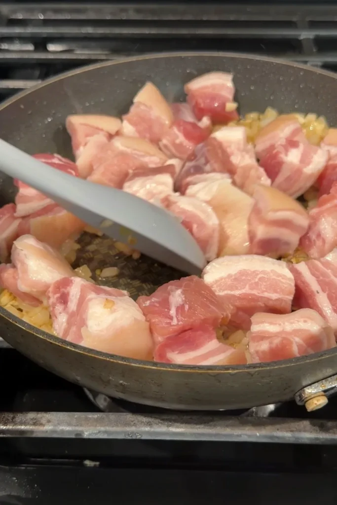 Raw pork belly cubes added to sautéed onion and garlic in a skillet, with a white spoon beginning to stir.