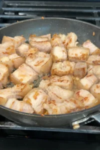Pork belly pieces browning in the pan with onion and garlic, with rendered fat starting to collect at the bottom.