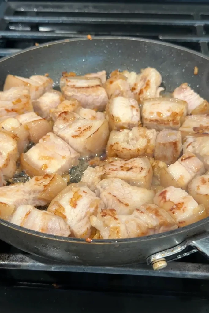 Pork belly pieces browning in the pan with onion and garlic, with rendered fat starting to collect at the bottom.
