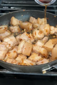 Fish sauce being poured into the skillet over lightly browned pork belly cubes.