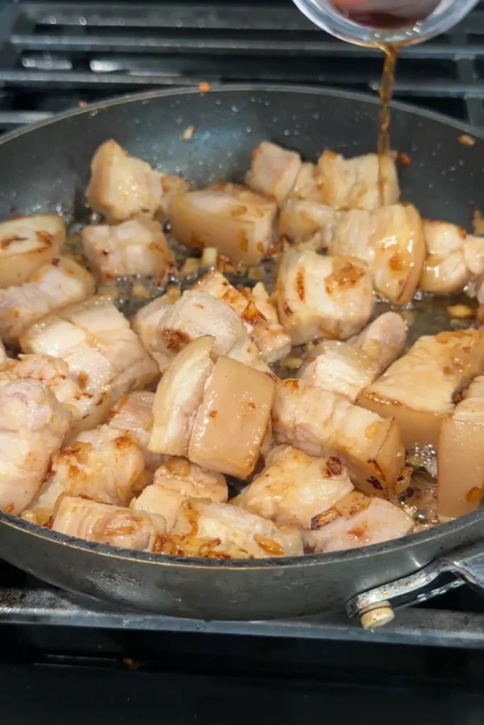 Fish sauce being poured into the skillet over lightly browned pork belly cubes.