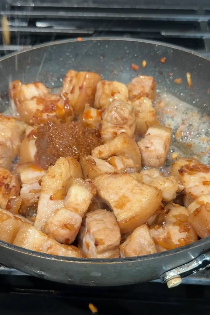 A spoonful of shrimp paste being added to browned pork belly in a sizzling skillet.