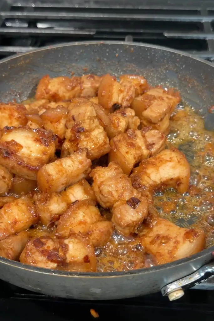Pork belly cooking with shrimp paste until coated and lightly caramelized, with oil bubbling around the edges.