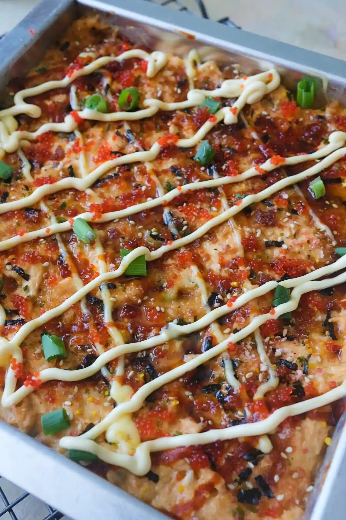 Close-up of baked salmon sushi in a rectangular metal pan, topped with zigzags of Kewpie mayo, red tobiko, chopped green onions, furikake, and glossy eel sauce over a lightly browned salmon layer.