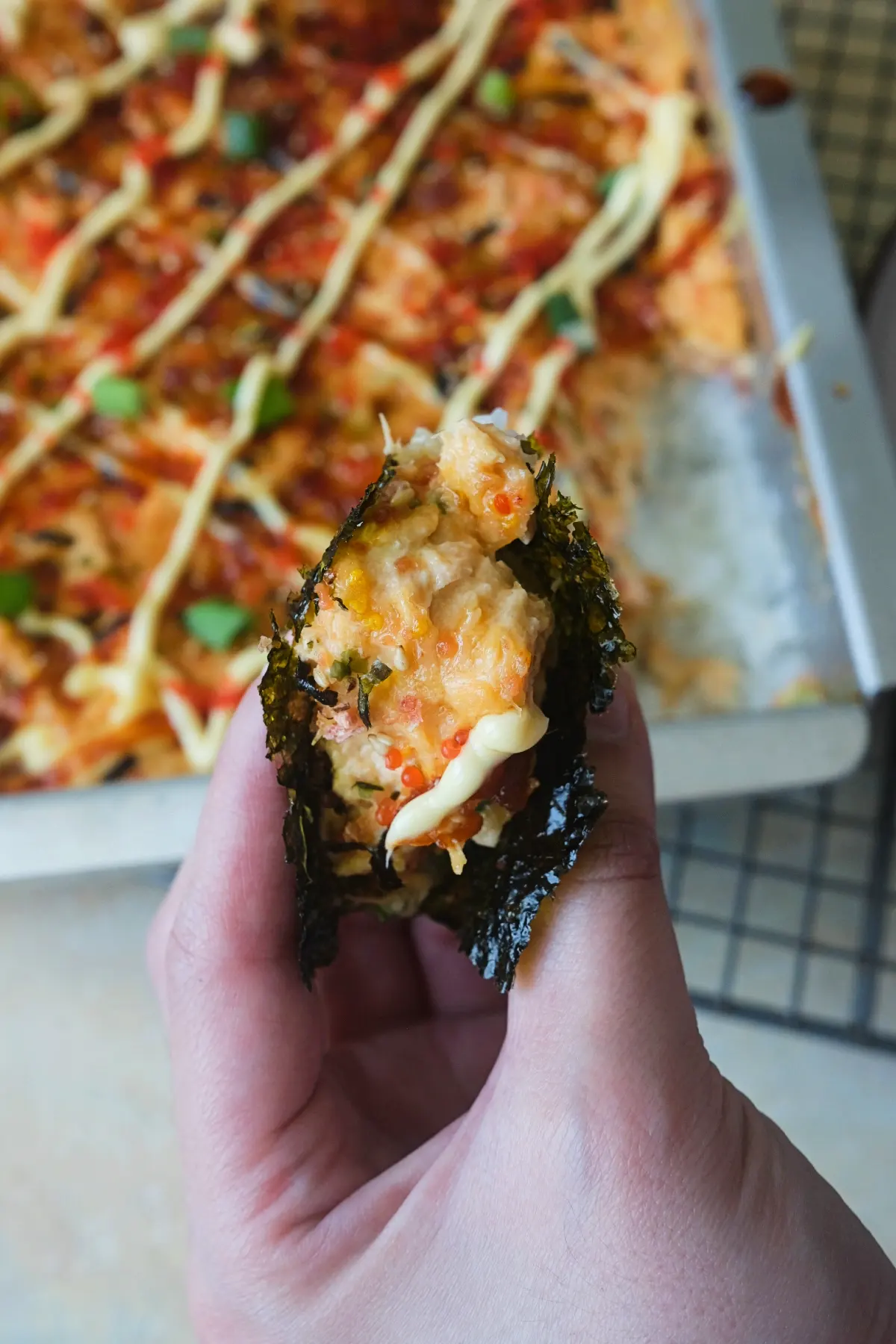 A hand holds a bite of salmon sushi bake wrapped in a sheet of roasted nori, showing creamy salmon, rice, tobiko, and sauce, with the pan of sushi bake blurred in the background.