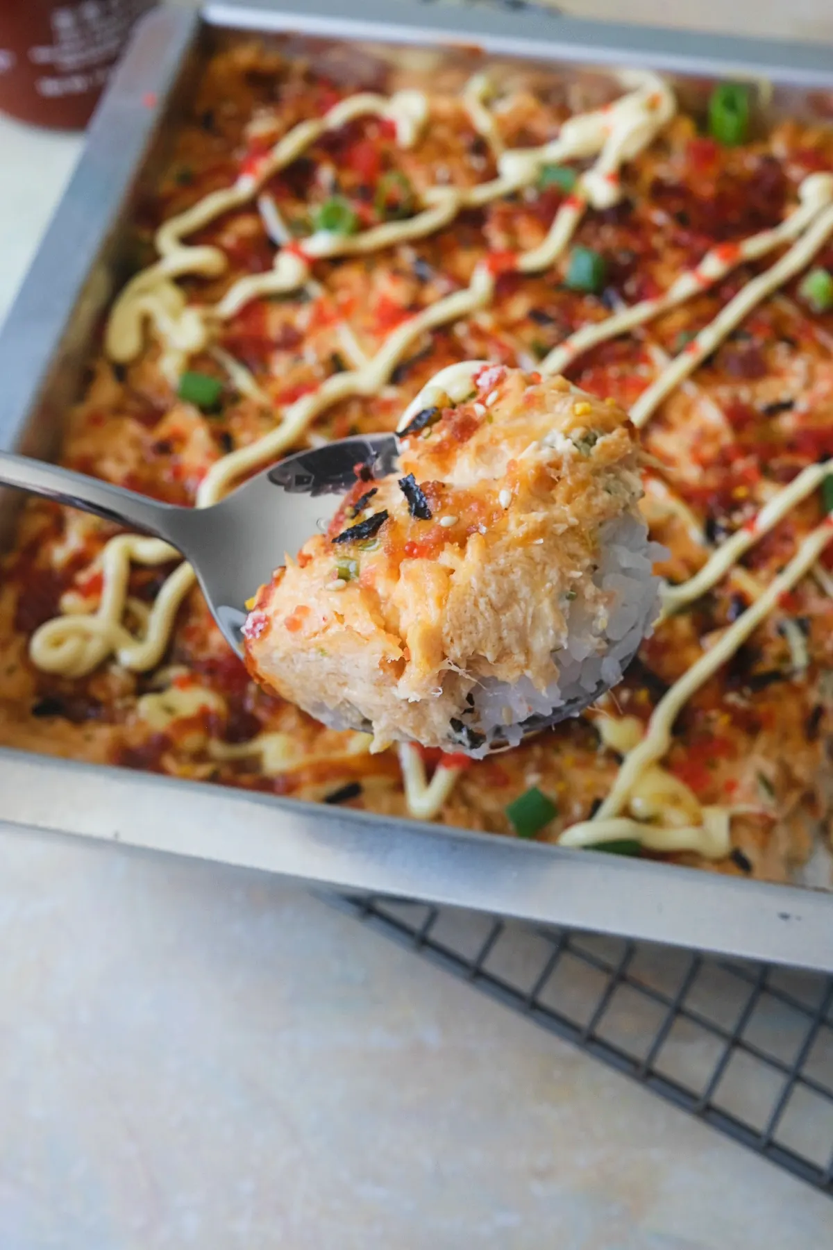 A spoon lifts a thick scoop of salmon sushi bake with layers of white sushi rice and creamy salmon filling, topped with furikake and tobiko, with the baking pan behind it.