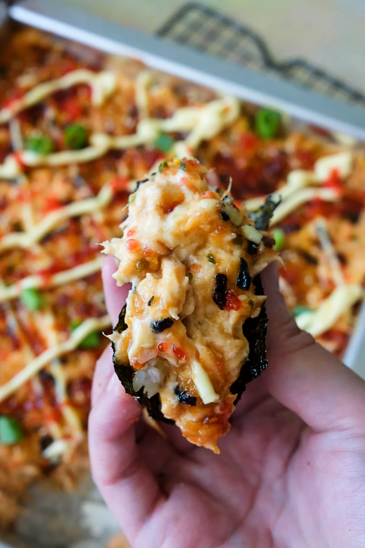 Close-up of a hand holding a loaded bite of salmon sushi bake in roasted nori, showing creamy salmon, eel sauce, furikake, and tobiko, with the rest of the bake softly blurred behind it.