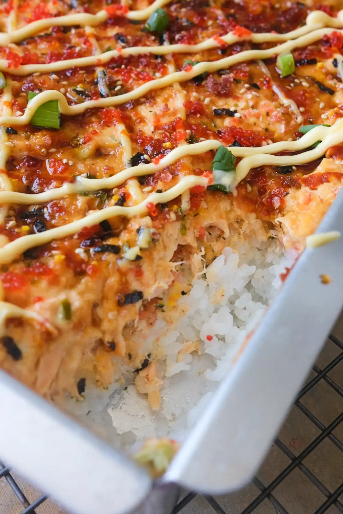 Close-up side view of salmon sushi bake in a metal pan, showing the thick layer of sticky white sushi rice and the creamy salmon topping