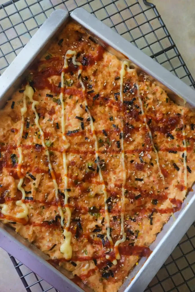 Overhead view of a rectangular pan of salmon sushi bake on a wire cooling rack, topped with drizzled Kewpie mayo, sriracha, and furikake