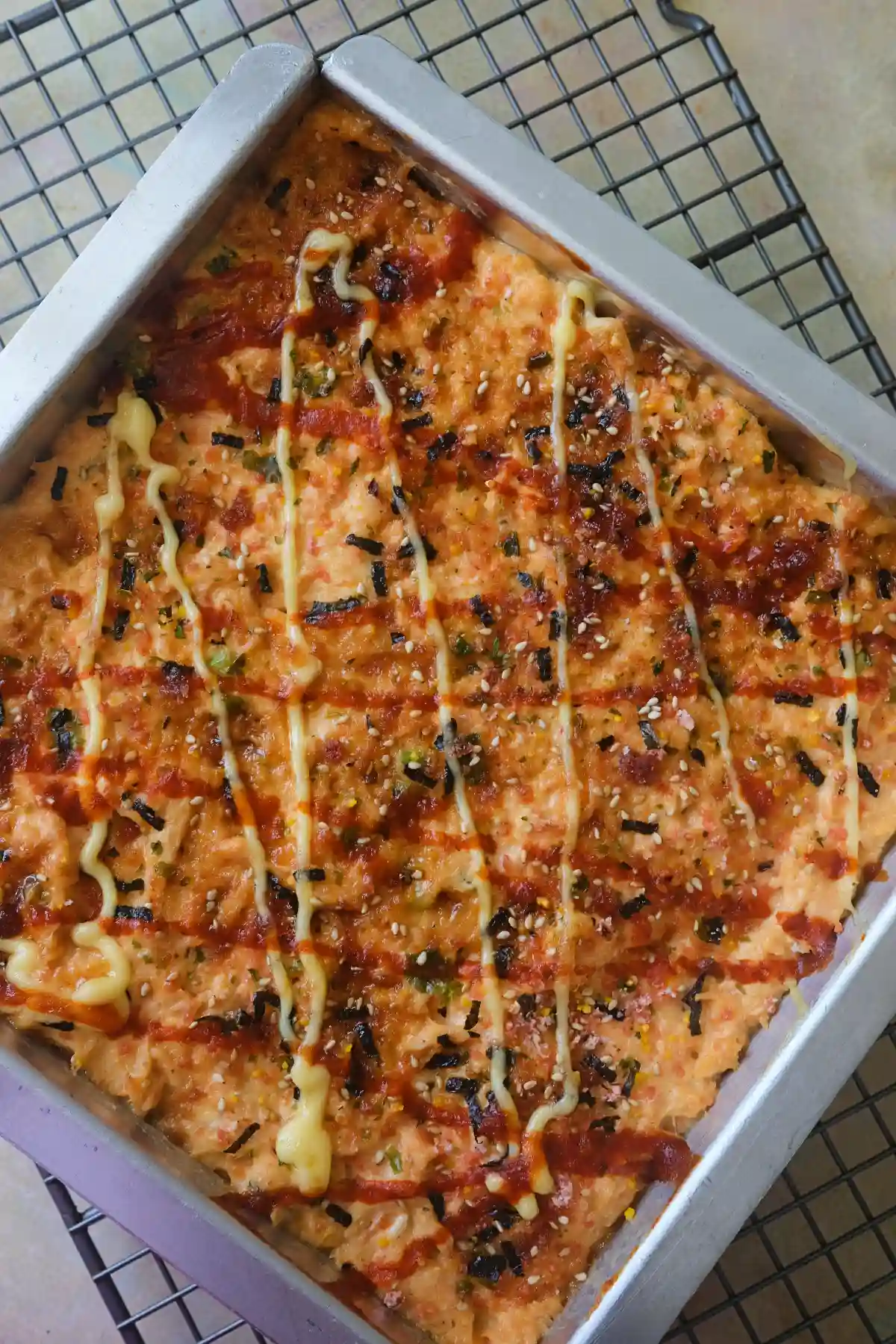 Overhead view of a rectangular pan of salmon sushi bake on a wire cooling rack, topped with drizzled Kewpie mayo, sriracha, and furikake