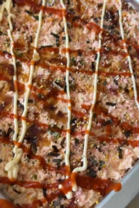 Unbaked salmon sushi bake in a metal pan is topped with drizzles of Kewpie mayo and sriracha, plus furikake and sesame seeds scattered across the surface.