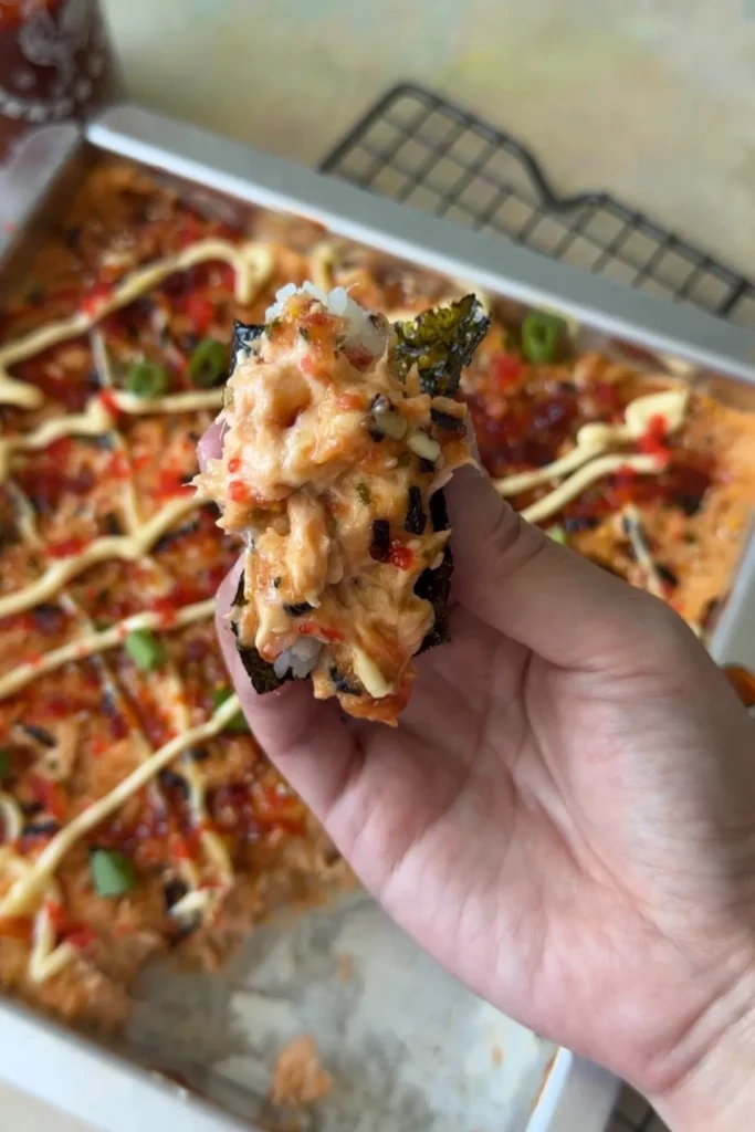 A hand holds a bite of salmon sushi bake wrapped in roasted nori, showing layers of rice and creamy salmon filling with tobiko and furikake, while the pan sits blurred in the background.