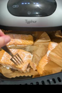 Fresh salmon fillets cook in a parchment-lined air fryer basket, while a fork presses into one piece to check doneness and flakiness.