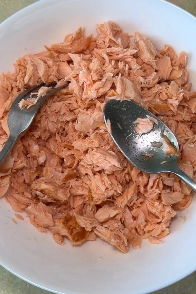 Cooked salmon flakes sit in a large white bowl with a fork and spoon, showing the tender texture after being broken into bite-sized pieces.