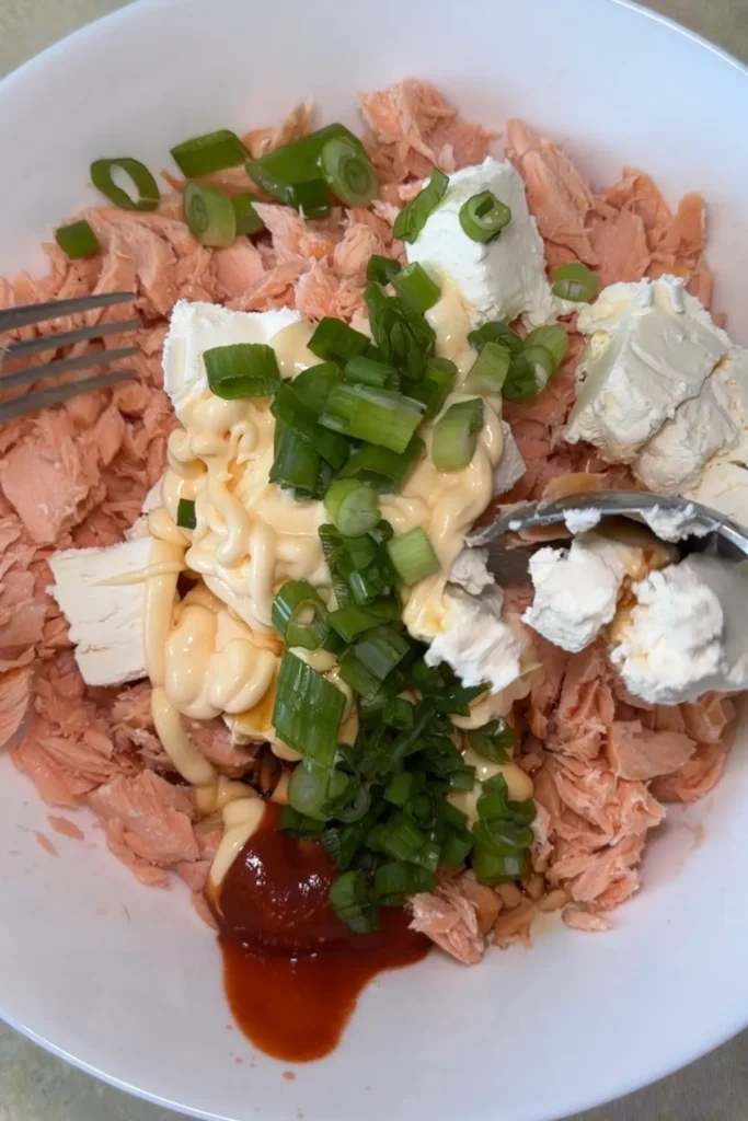 Flaked salmon in a white bowl is topped with chunks of cream cheese, Kewpie mayo, sriracha, and sliced green onions before being mixed into the filling.