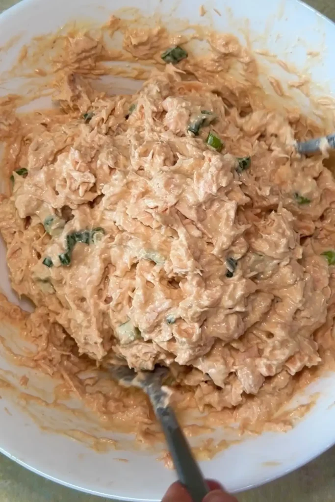 Creamy salmon mixture is stirred together in a white bowl until well combined, with green onion pieces visible throughout the pale orange filling.