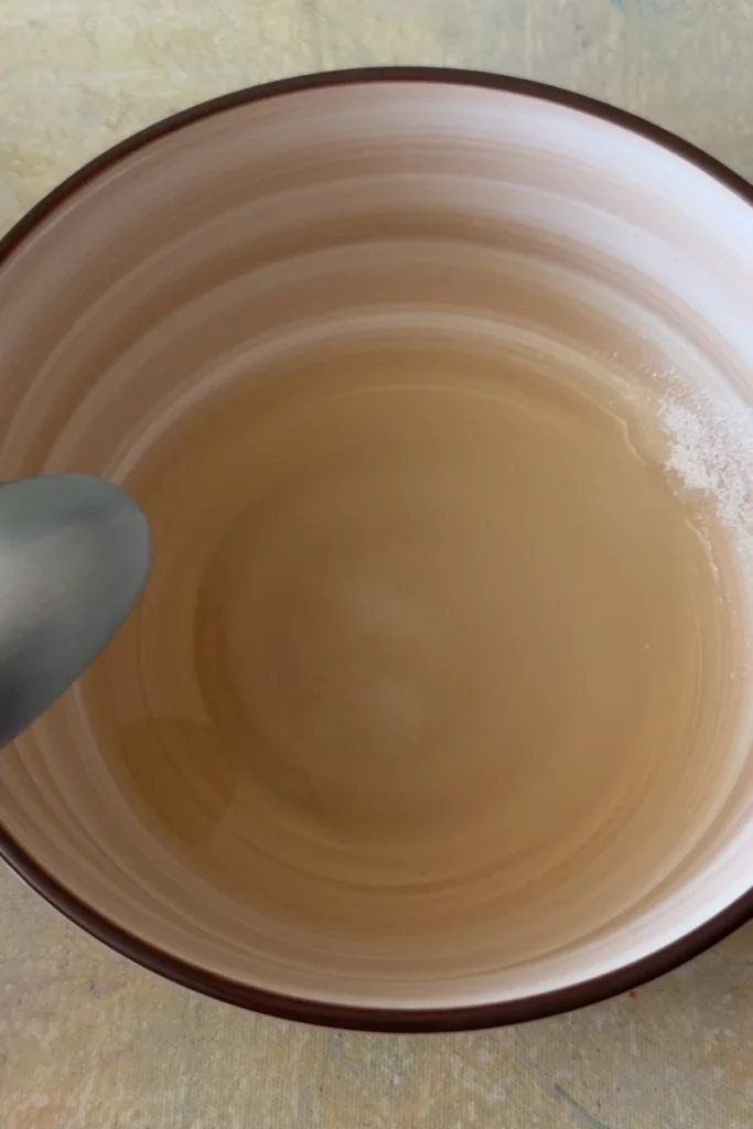Rice vinegar mixture for sushi rice sits in a tan bowl with sugar still visible along the edge before being stirred to dissolve.
