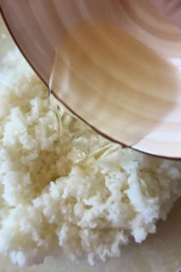 Sushi rice seasoning is poured from a tan bowl over freshly cooked white rice.
